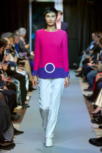 Model walks the runway in a hot-pink top with purple hem and circular accents, paired with light blue glossy pants.
