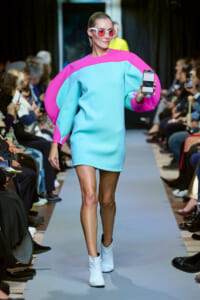Model walks a fashion show runway in a bright turquoise mini dress with pink cape sleeves, white sunglasses, and white ankle boots, holding a smartphone toward the camera.