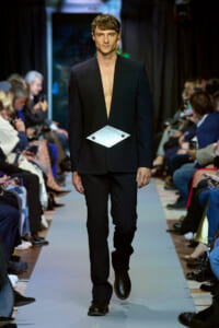 Male model walking a fashion runway in a black deep-plunge blazer with a diamond-shaped silver belt buckle over the midsection.