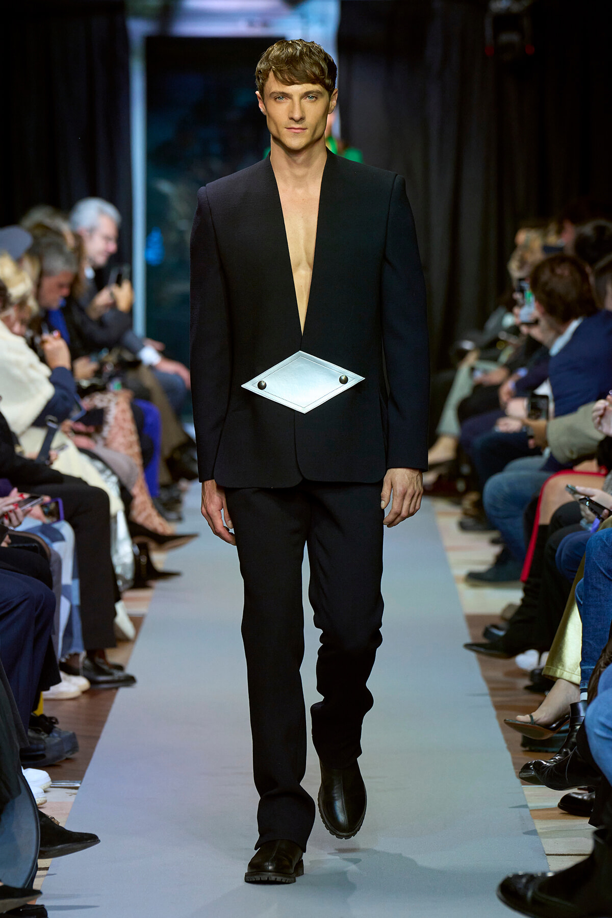 Male model walking a fashion runway in a black deep-plunge blazer with a diamond-shaped silver belt buckle over the midsection.