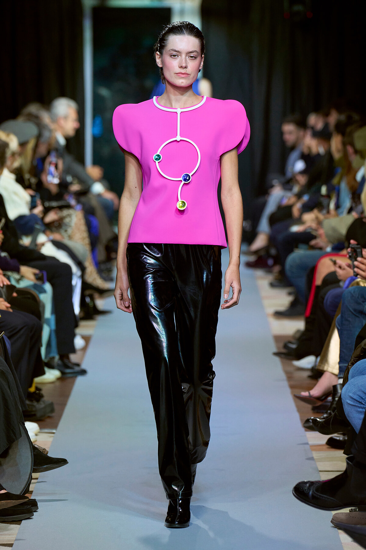Model walks the runway in a vivid pink sculptural top with white piping and circular gemstone accents, paired with glossy black pants.