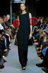 Model walks the runway in a sleeveless black gown with oversized red cage-like spheres at the hips and a confident smile on her face.