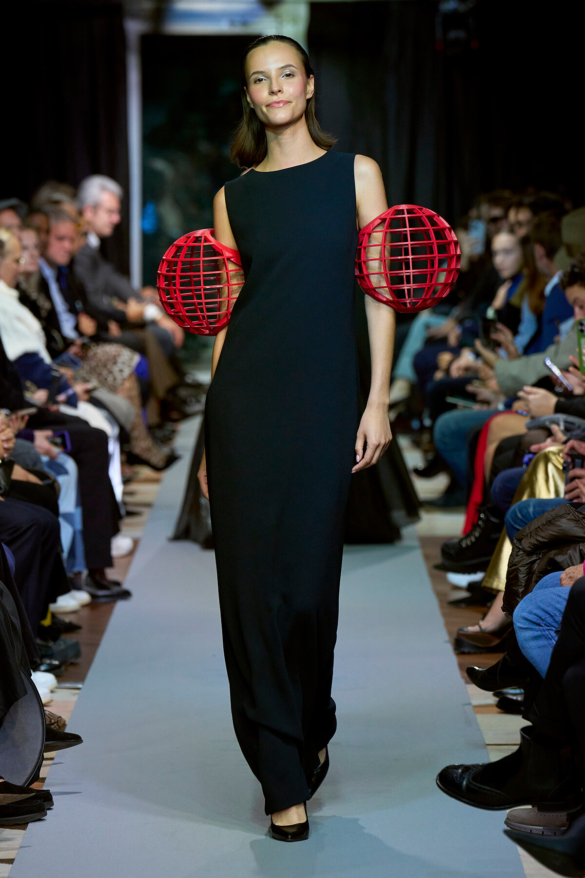 Model walks the runway in a sleeveless black gown with oversized red cage-like spheres at the hips and a confident smile on her face.
