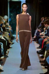 Model walks a fashion runway in a sleeveless brown jumpsuit with a beige waist stripe, audience seated on both sides framing the catwalk.