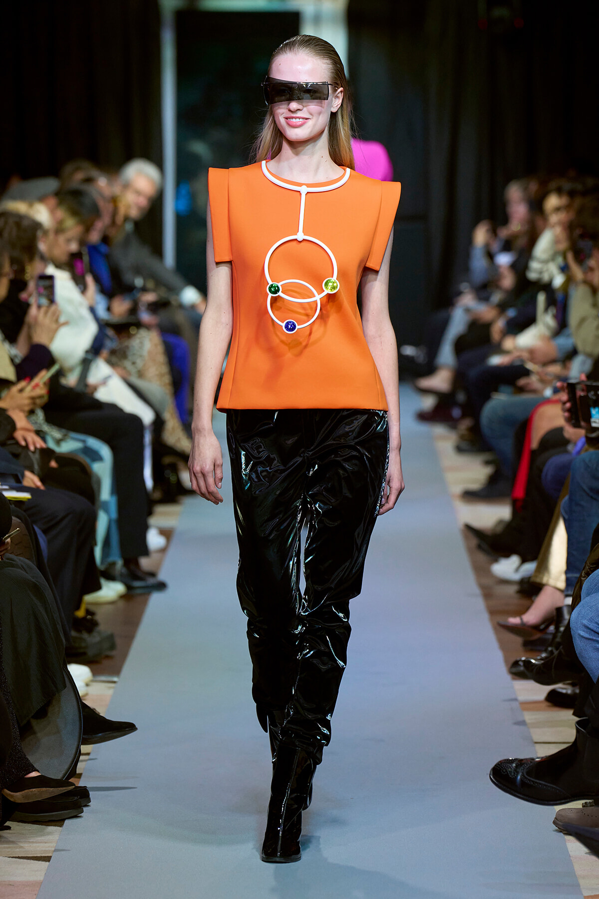 Model walking a runway in an oversized orange top with white geometric necklace and glossy black pants; futuristic sunglasses on a crowded show stage