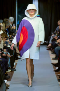 Model walks the runway in a pale white oversized hooded coat with a bold purple, red, and orange color-block panel on the front.