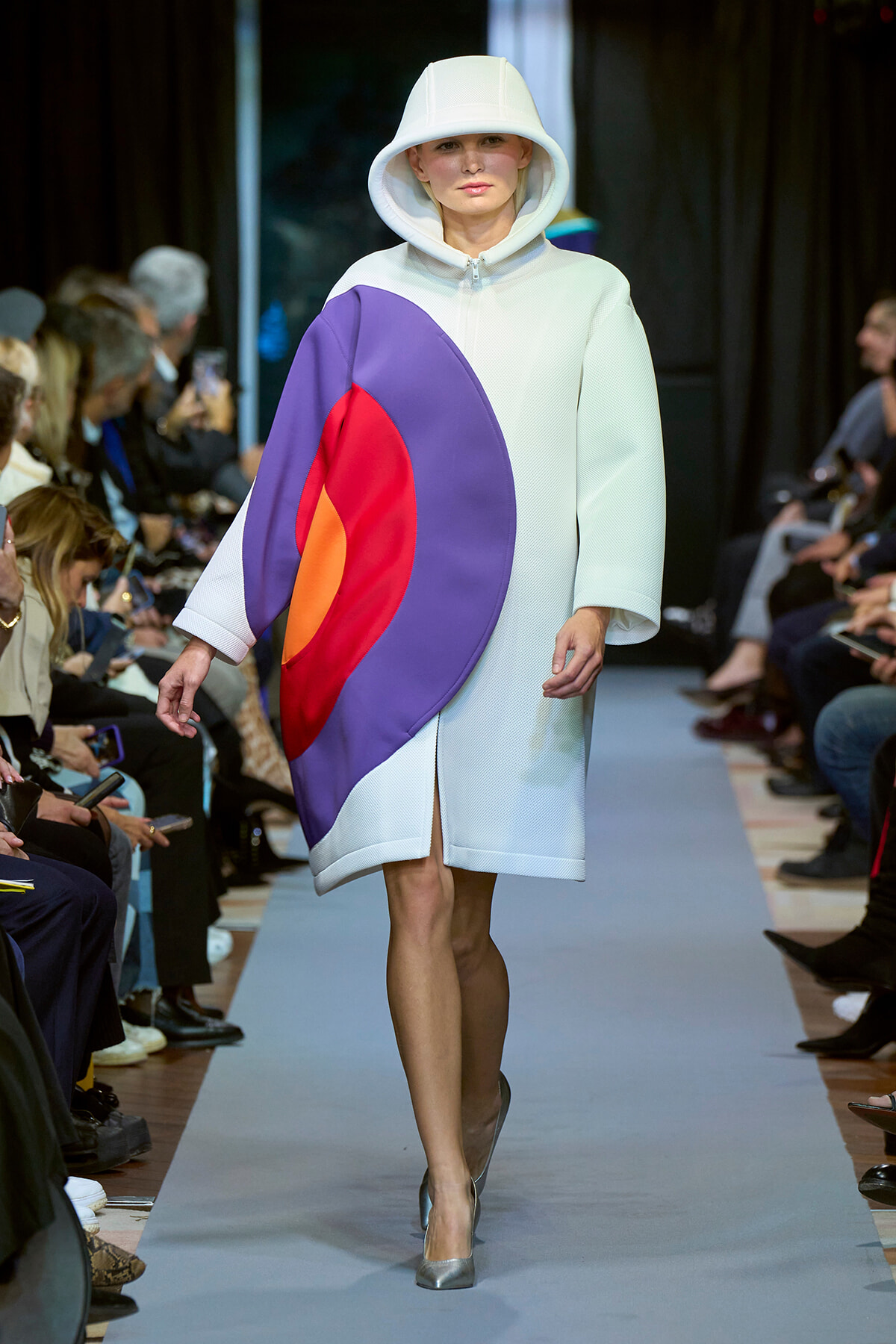 Model walks the runway in a pale white oversized hooded coat with a bold purple, red, and orange color-block panel on the front.