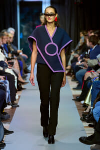 Model walks the runway in a navy poncho with lilac trim and a circular motif, paired with black pants and red earrings, wearing sunglasses.