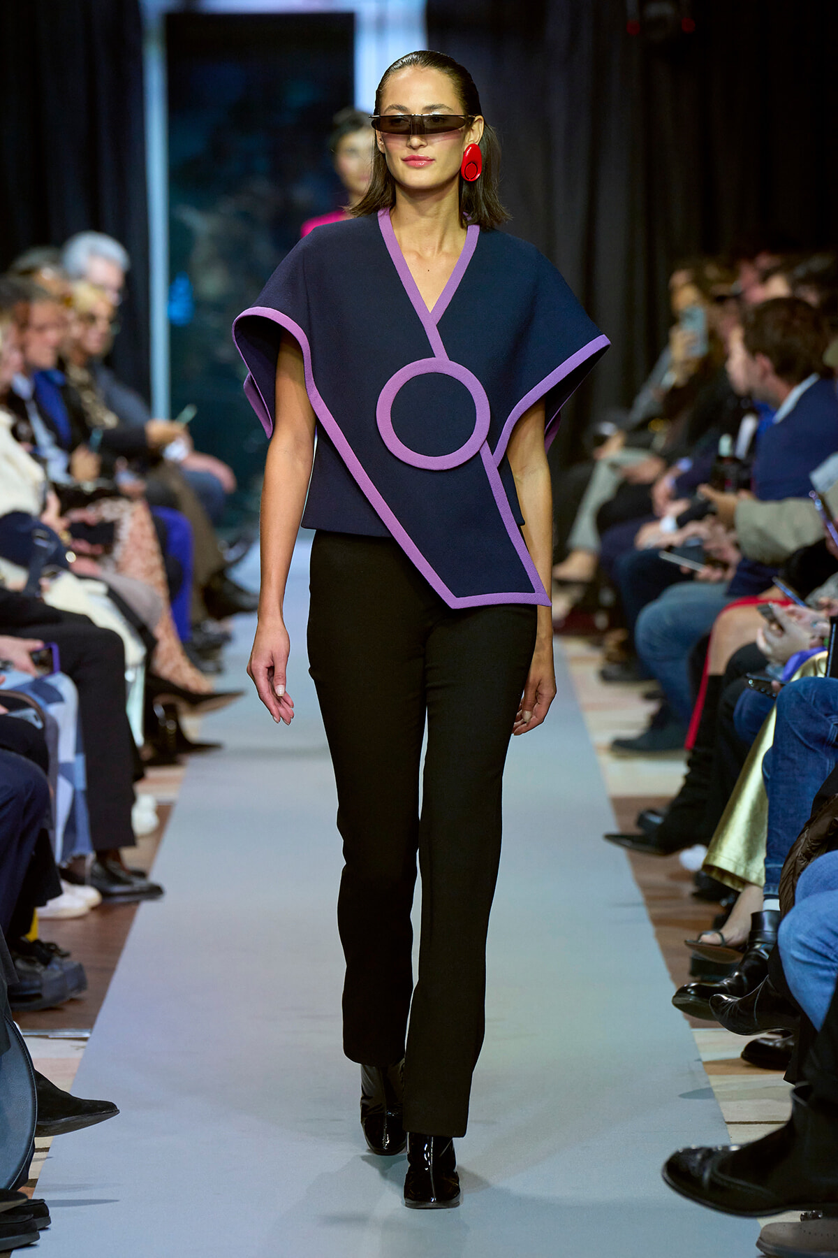 Model walks the runway in a navy poncho with lilac trim and a circular motif, paired with black pants and red earrings, wearing sunglasses.