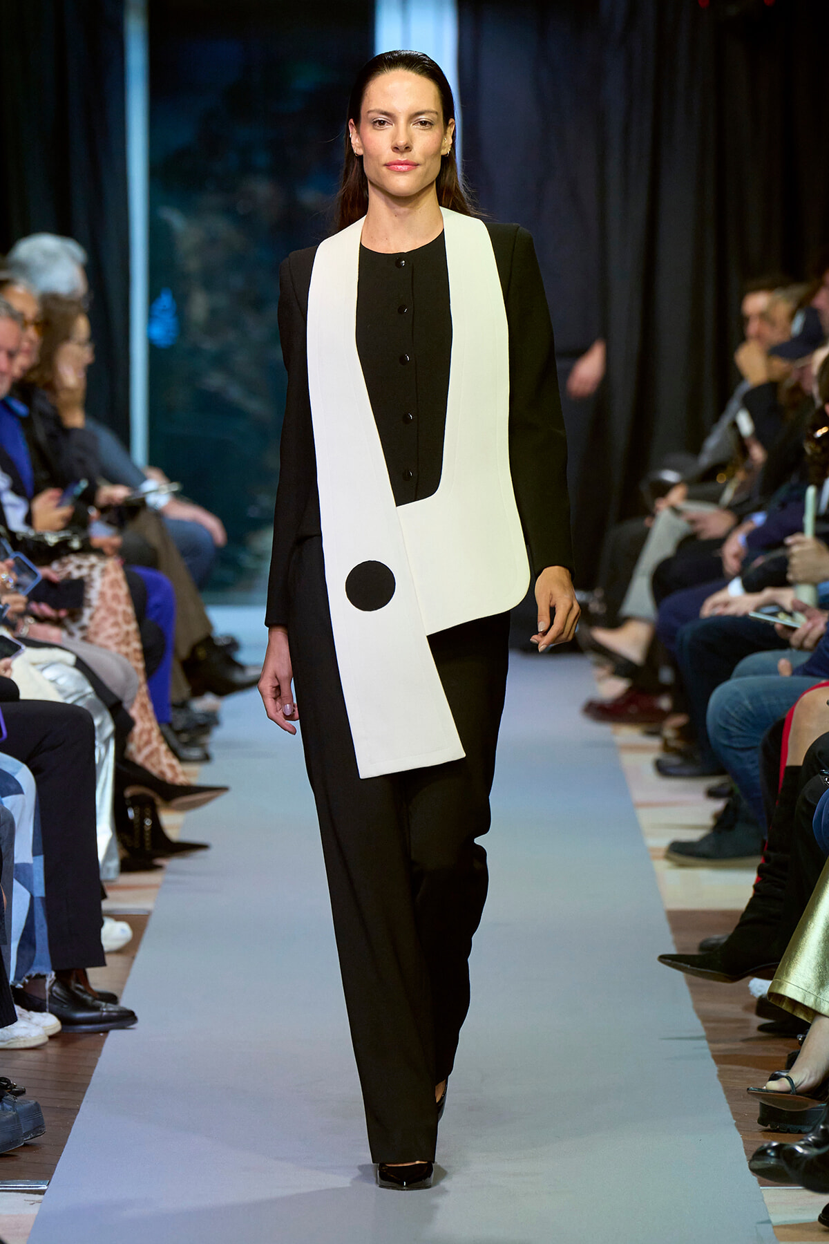 Model walking a fashion runway wearing a black suit with a long white sleeveless vest featuring a large black circle detail at the waist.