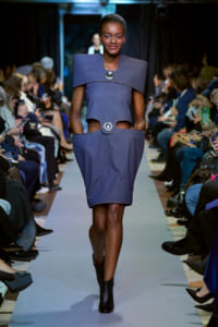 Model walks down a runway wearing a structured blue dress with oversized shoulder collar and circular metal accents, audience seated on both sides capturing photos.