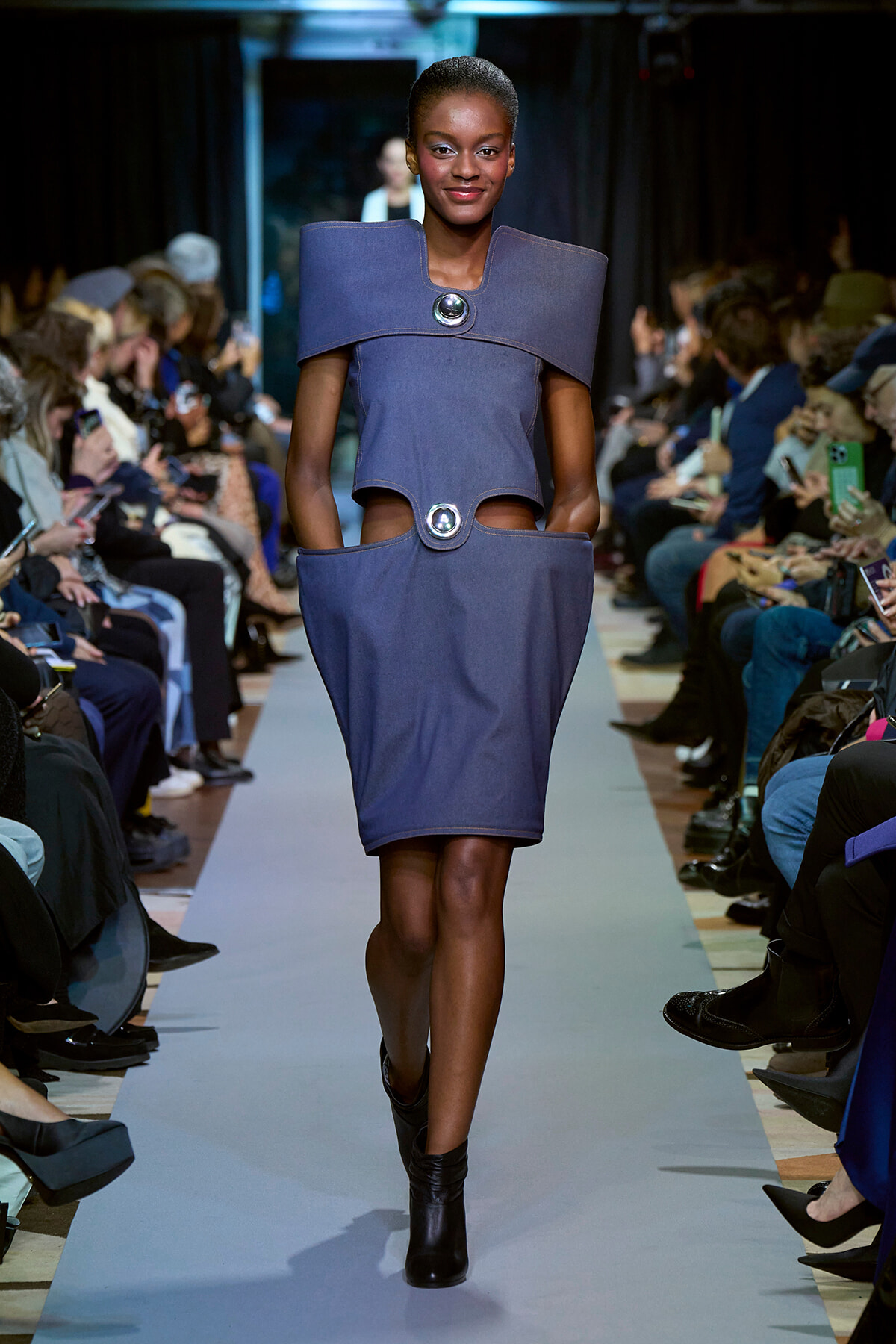 Model walks down a runway wearing a structured blue dress with oversized shoulder collar and circular metal accents, audience seated on both sides capturing photos.
