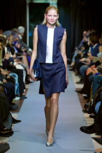 Model on runway wearing a navy sleeveless dress with a white center panel, carrying a blue clutch and wearing silver pumps.