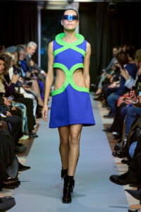 Model walks a blue and lime-green geometric mini dress on a runway, wearing futuristic visor sunglasses.