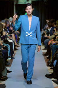 Male model walks the runway in a light-blue suit with a deep V-neck and a white cross-shaped clasp; audience blurred in the background.