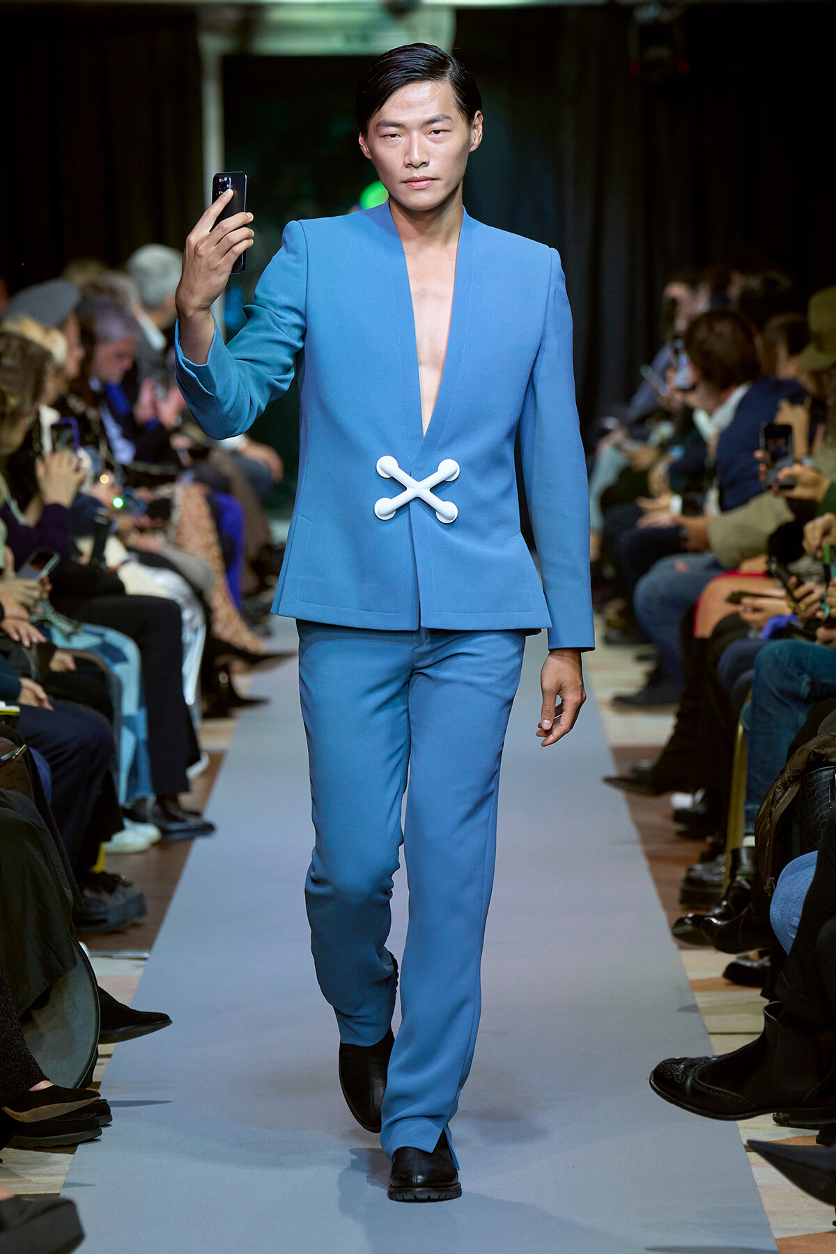 Male model walks the runway in a light-blue suit with a deep V-neck and a white cross-shaped clasp; audience blurred in the background.