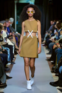 Model walks a fashion runway wearing a tan two-piece outfit with a white X-belt and white ankle boots, oversized white sunglasses with yellow lenses, and a curly hairstyle.