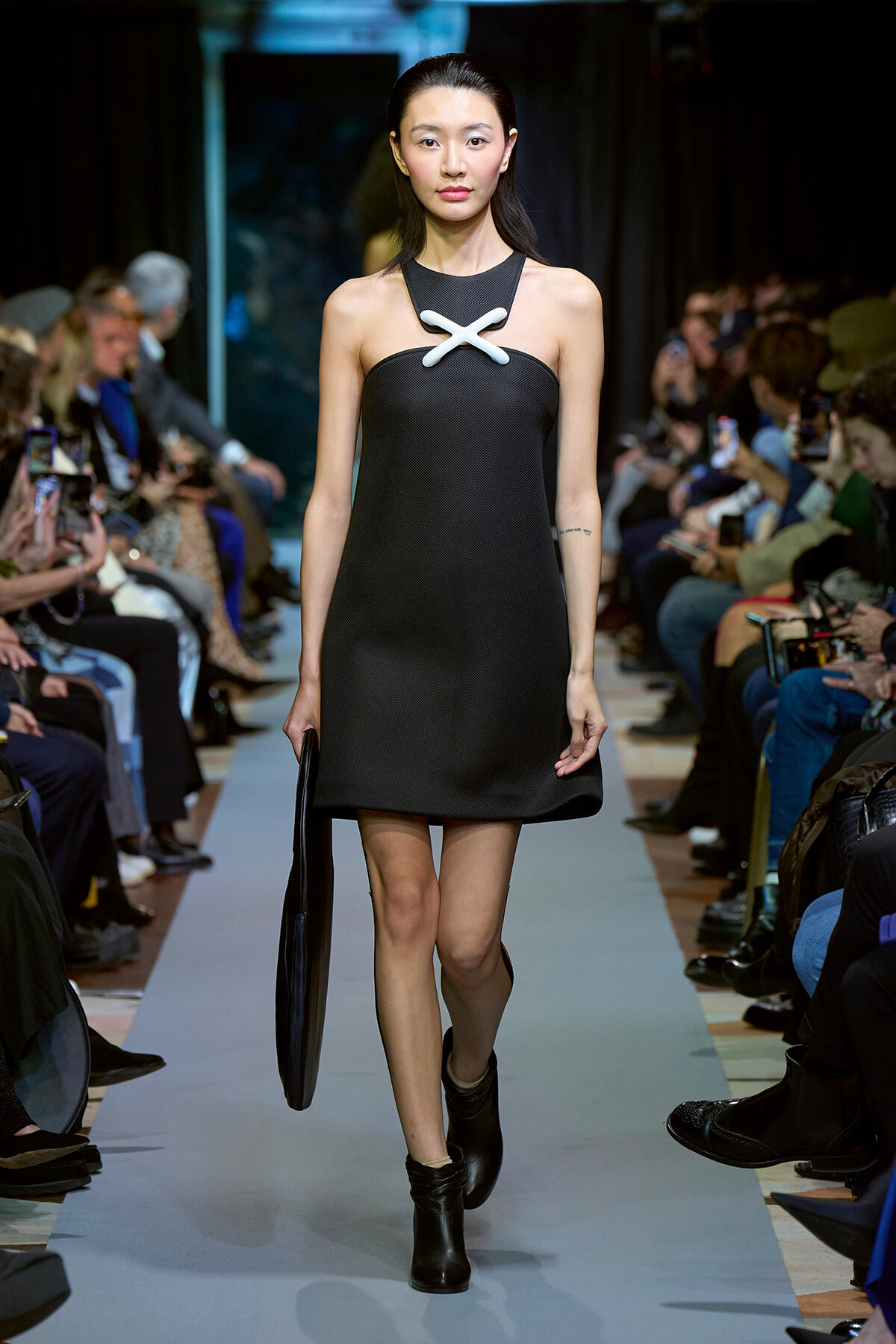 Model walks a runway in a black sleeveless shift dress with a white X-shaped neckline accent, carrying a black clutch.