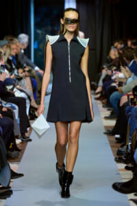 Model on a fashion runway wearing a black sleeveless mini dress with white sculpted shoulders, oversized visor sunglasses, black ankle boots, and a white geometric clutch.