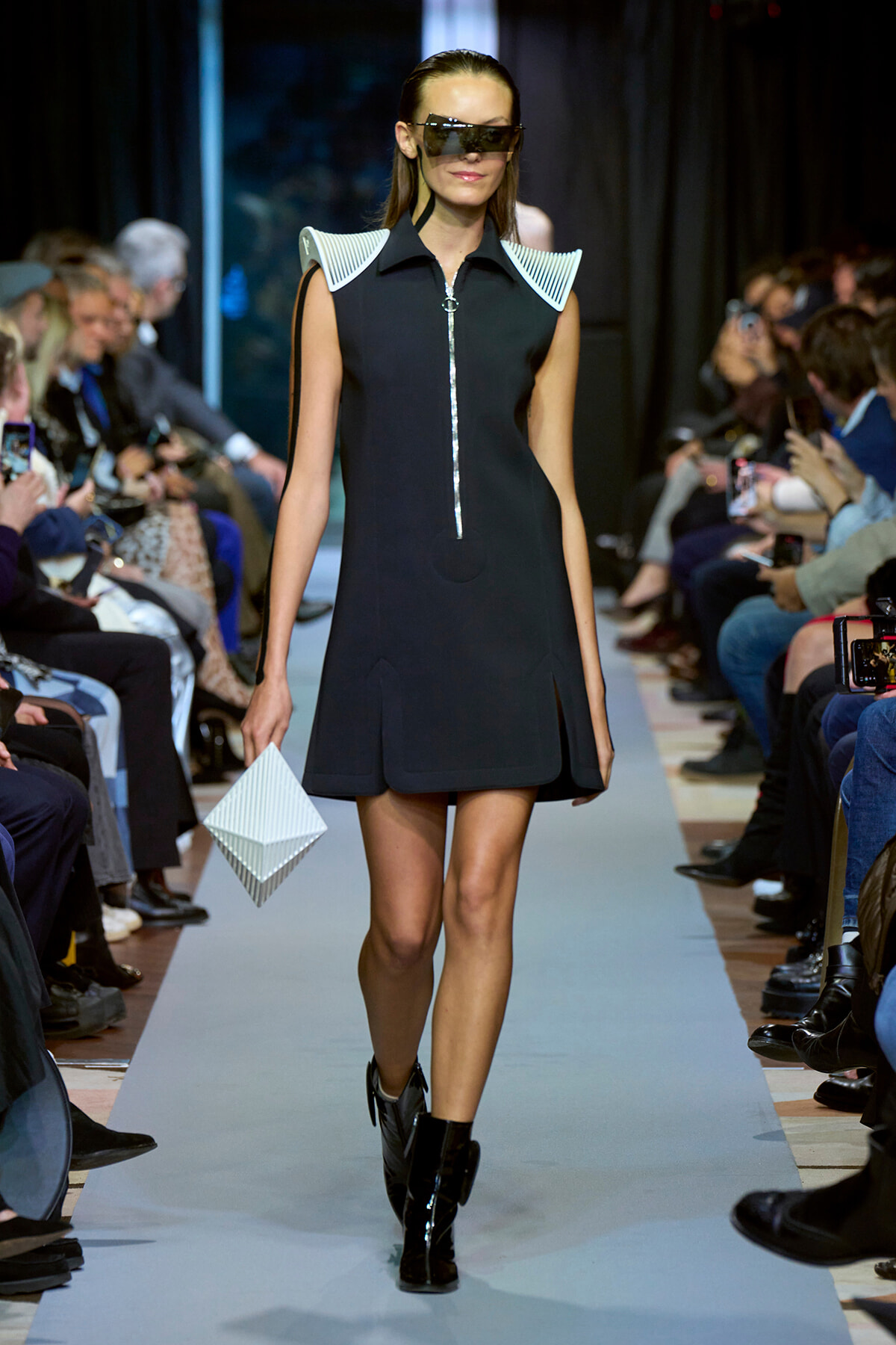 Model on a fashion runway wearing a black sleeveless mini dress with white sculpted shoulders, oversized visor sunglasses, black ankle boots, and a white geometric clutch.