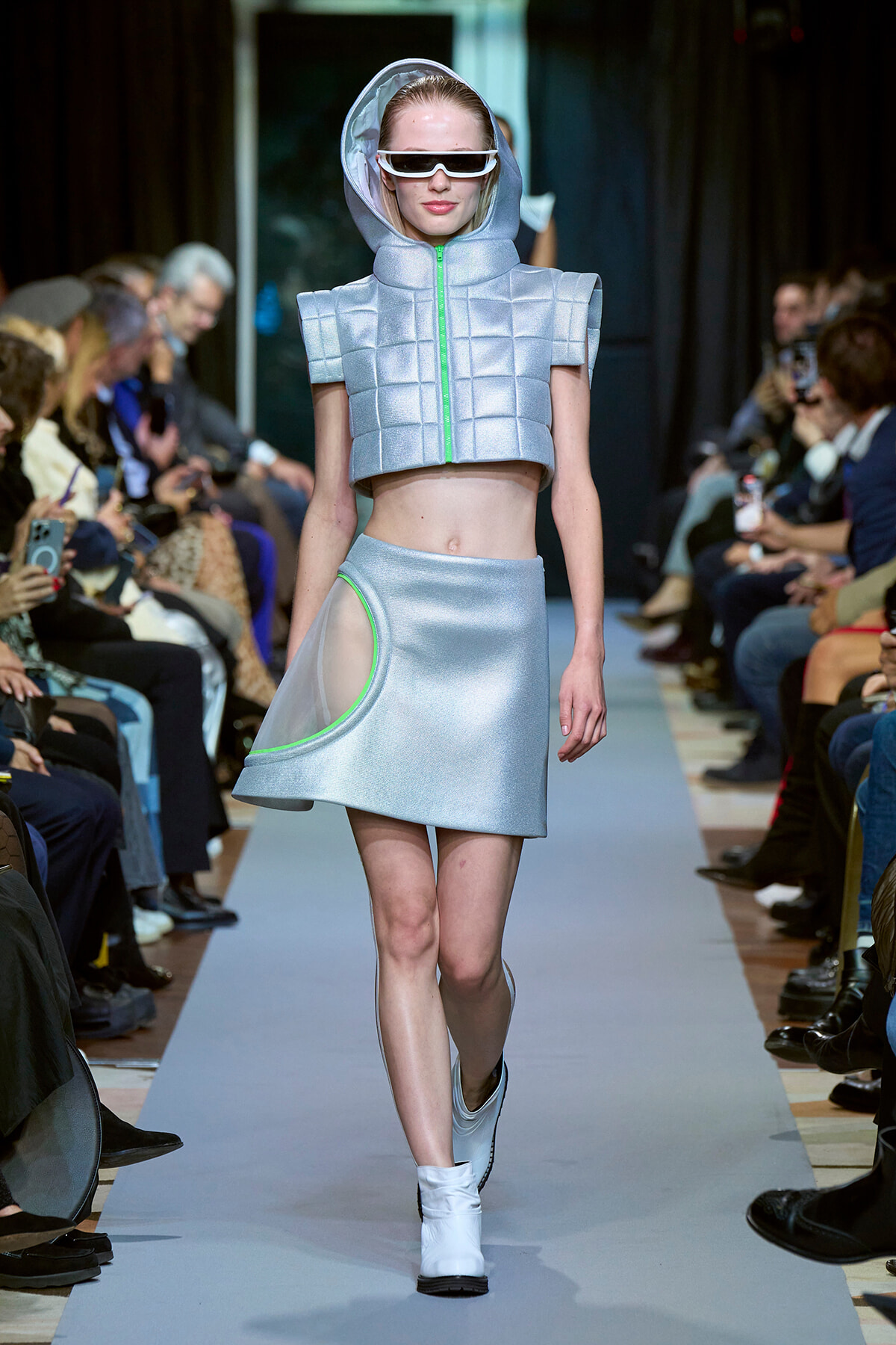 Model walks the runway in a silver padded crop jacket with hood, matching skirt with neon green trim and white visor sunglasses.