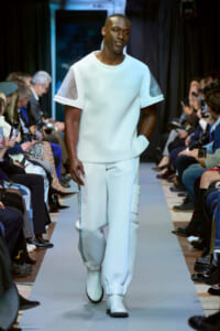 Male model walks a runway in a pale blue outfit with sheer sleeve panels; audience lines on both sides.