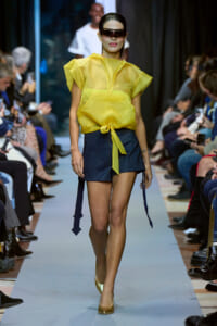 Model walking a runway in a bright yellow sheer top tied at the waist and navy shorts, wearing futuristic sunglasses and gold heels, audience on sides