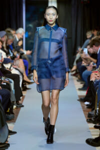 Model walks the runway in a sheer blue outfit with matching shorts and black ankle boots; audience sits along the catwalk.