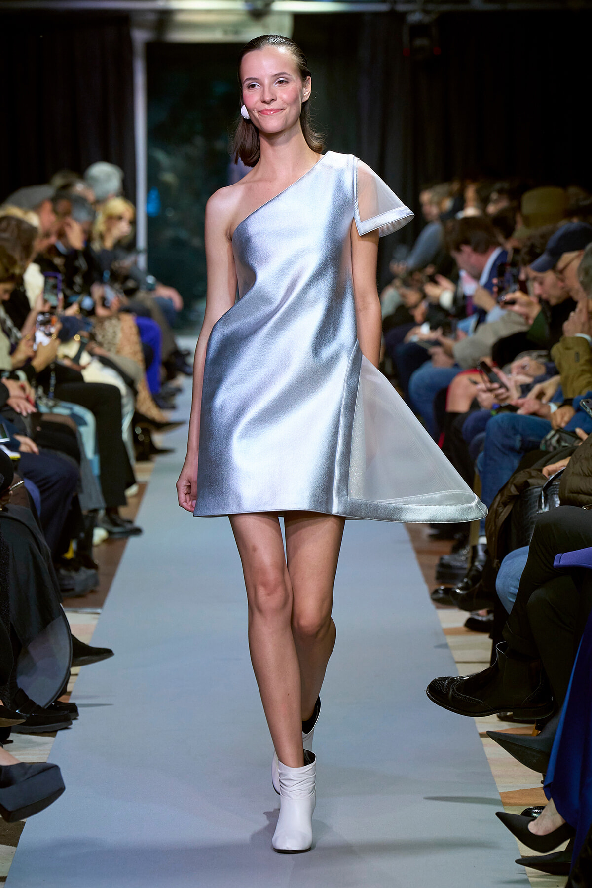 Model walks the runway in a silver one-shoulder mini dress with a flowing vent on the side, audiences on both sides.
