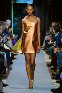Female model walks a fashion runway in a copper metallic strapless mini dress, with a yellow-green panel peeking from the hem and gold heels.