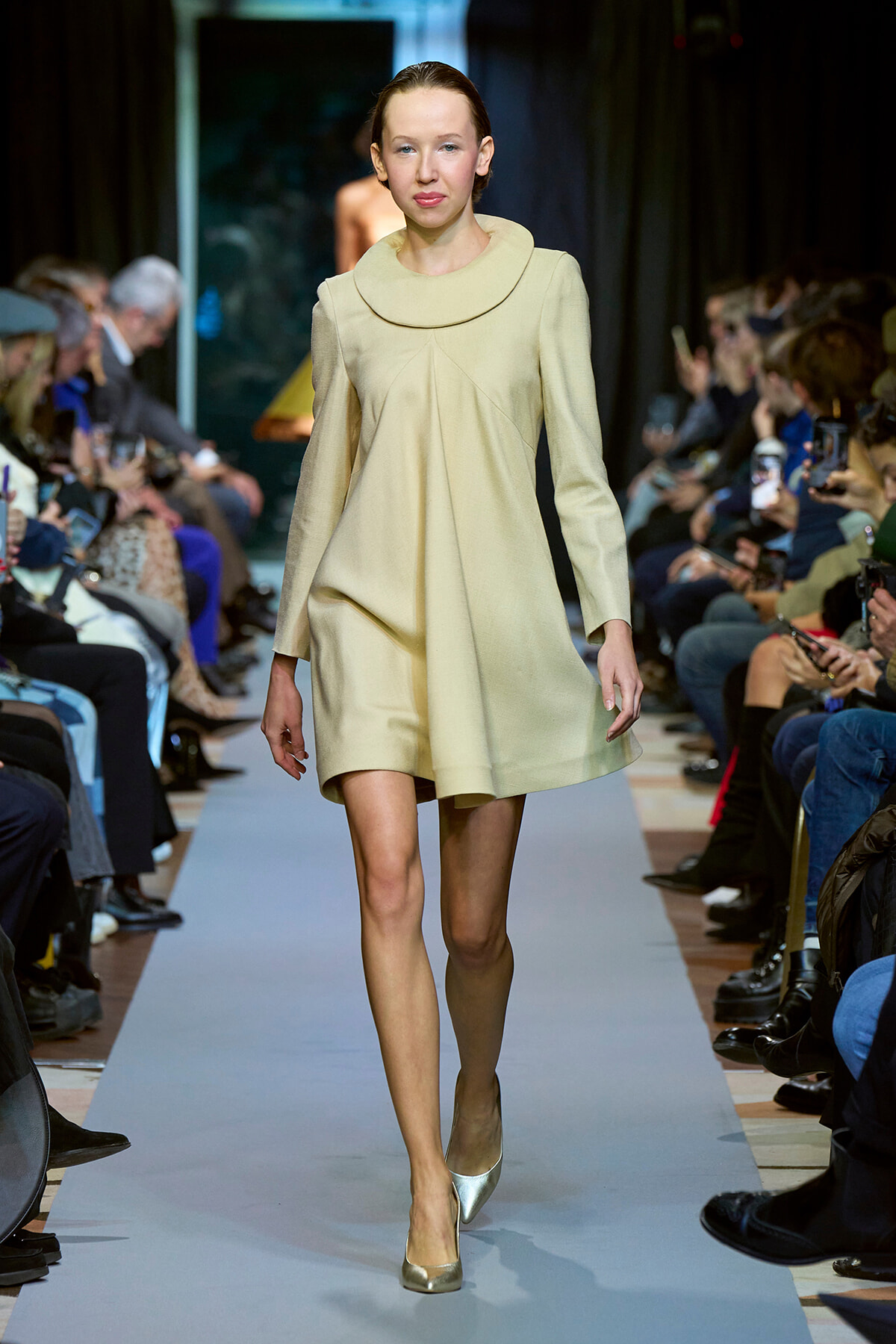 Model walks the runway in a pale beige A-line dress with a large rounded collar; audience watches from both sides.