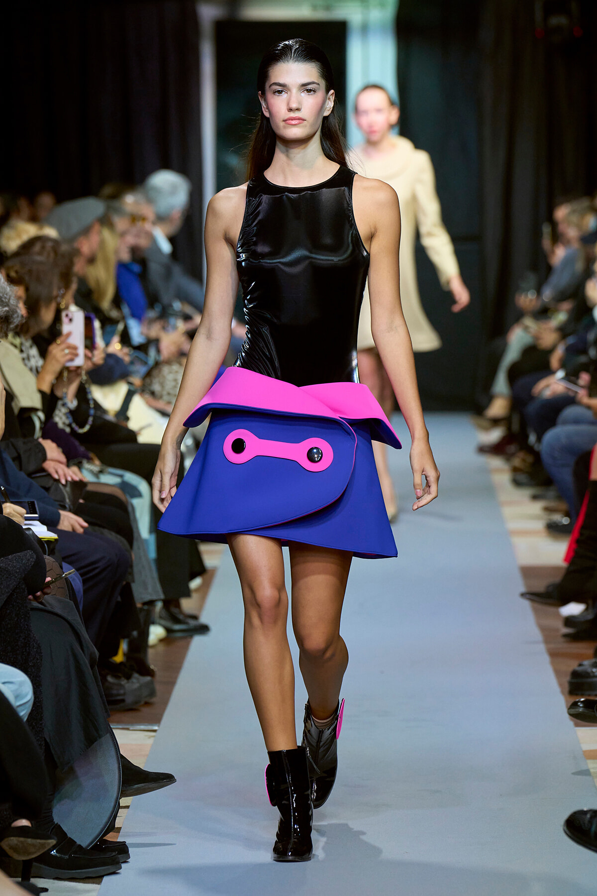 Model walks a runway wearing a glossy black sleeveless top and a bold blue and hot-pink sculptural skirt with a pink waist fold and oversized pocket detail in front.