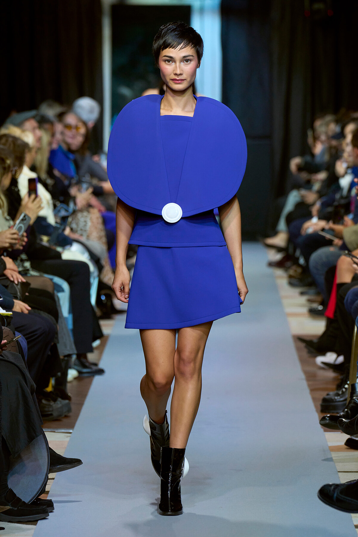 Model walks a runway in a cobalt blue sculptural dress with oversized circular shoulder cape and a white belt detail. Boots visible beneath the hem.