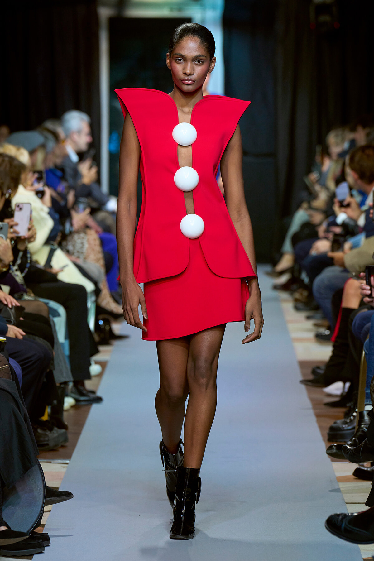 Model walks the runway in a red sculptural dress with three large white spheres down a sheer center panel, black boots visible.
