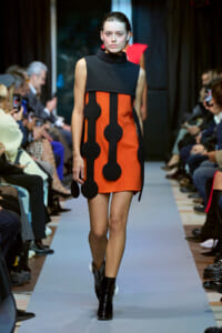 Model walks the runway in an orange dress with black, circular appliqués and a black sleeveless top; audience seated along the sides.