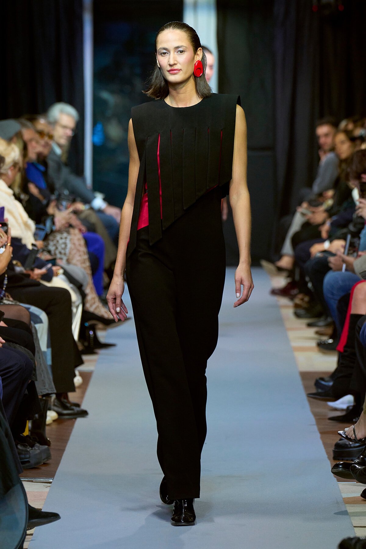 Model walking a fashion runway in a black sleeveless jumpsuit with red accents and red earrings, audience on both sides watching