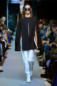 Model walks the runway in a black cape with vertical slit panels over pale blue pants and white boots, wearing oversized sunglasses.