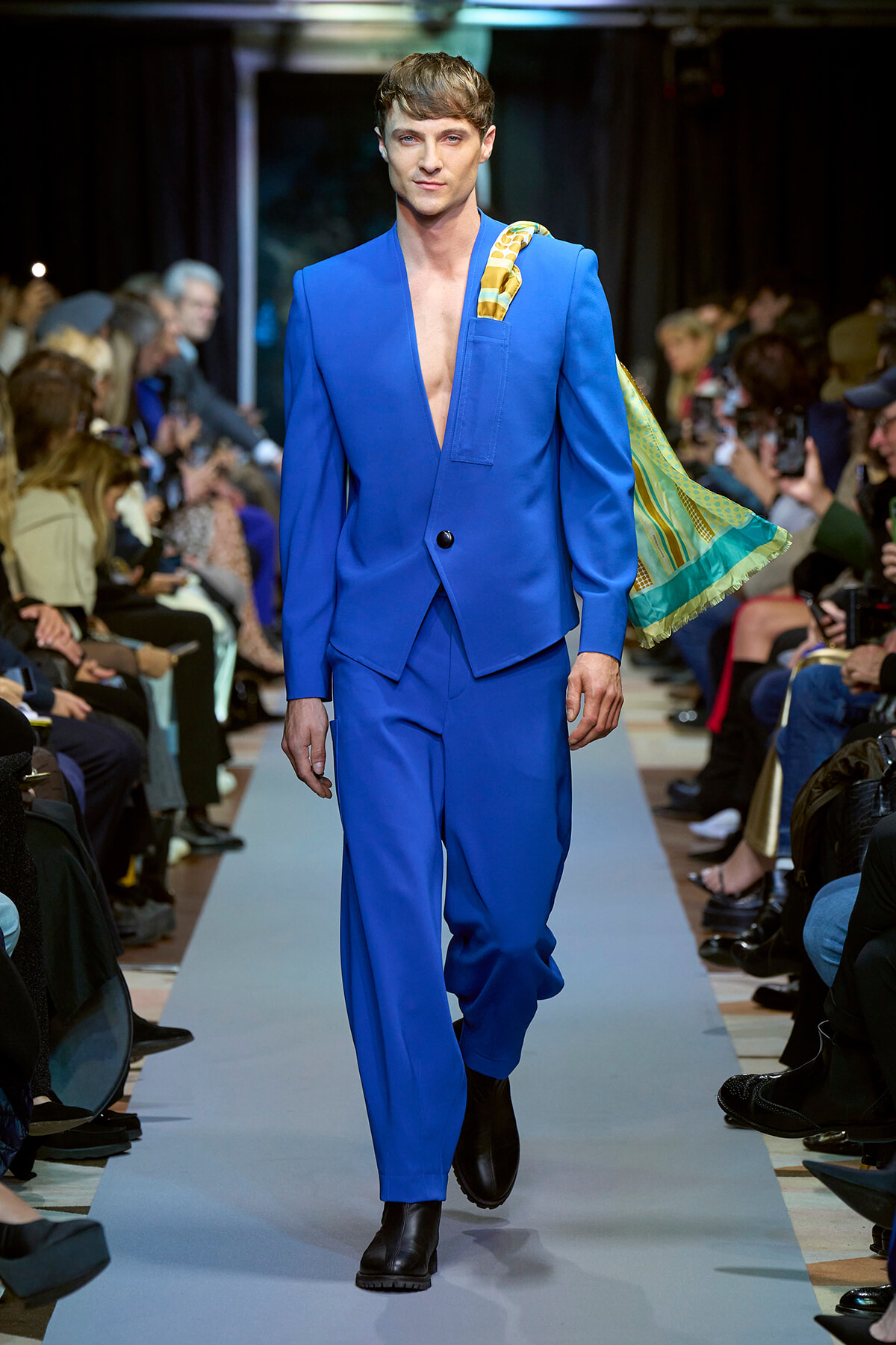 Male model in a bright cobalt blue suit walks the runway during a fashion show, with a striped scarf draped over his shoulder and spectators in the background.
