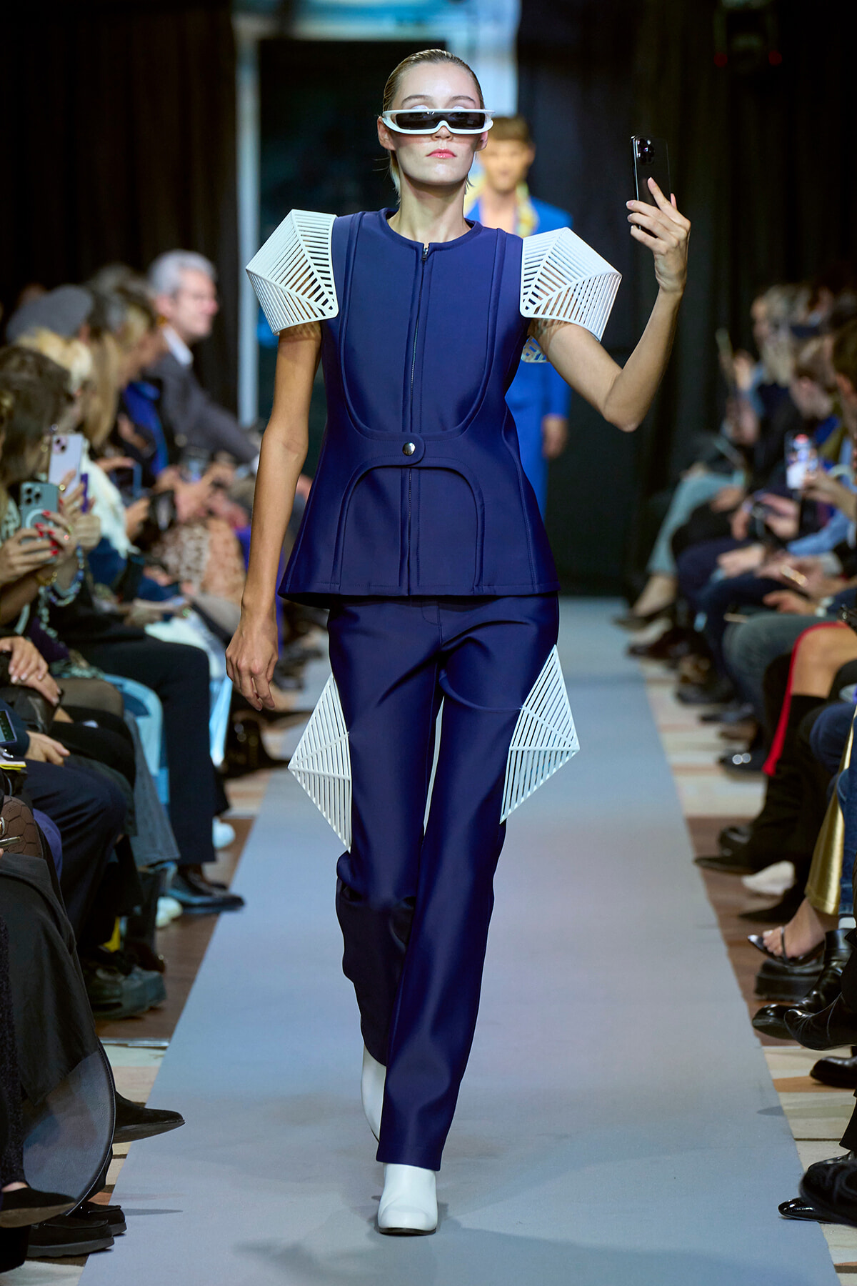 Model on a runway in a navy blue, futuristic outfit with white geometric shoulder and hip panels, wearing white sunglasses and taking a selfie with her phone.