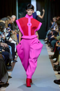 Model walks a runway in a bold hot-pink sculptural outfit with bright pink trim and red ankle boots, audience on both sides.