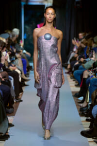 Model walks the runway in a metallic lavender strapless gown with a round blue gem at the bust.