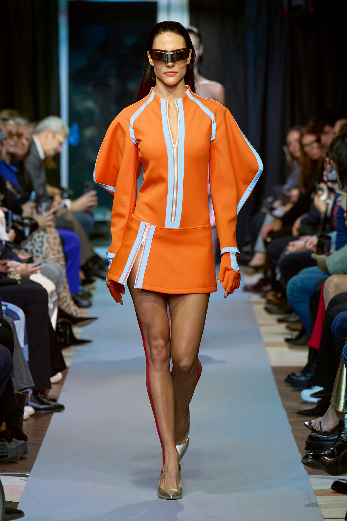 Model walks runway in bright orange futuristic dress with light blue trim and oversized sunglasses.
