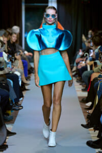 Model walking a fashion runway in a metallic blue sculptural top with oversized shoulders and matching turquoise mini skirt, white ankle boots, and round sunglasses.