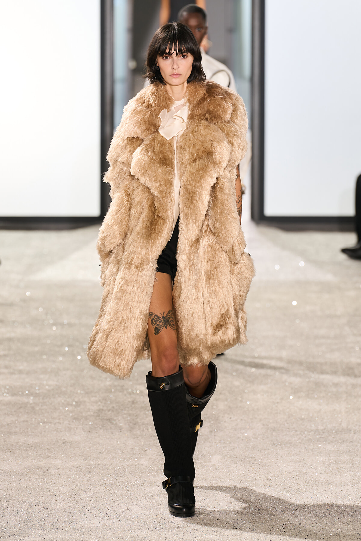 Model walks the runway in a large beige faux-fur coat over a pale blouse, black shorts, and knee-high black boots; visible thigh tattoo.