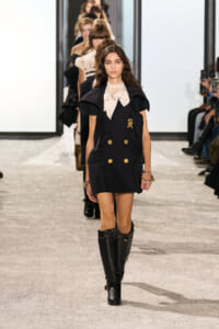 Model walks the runway in a navy double-breasted coat-dress with gold buttons, beige blouse, and knee-high black boots, as other models line up behind her.