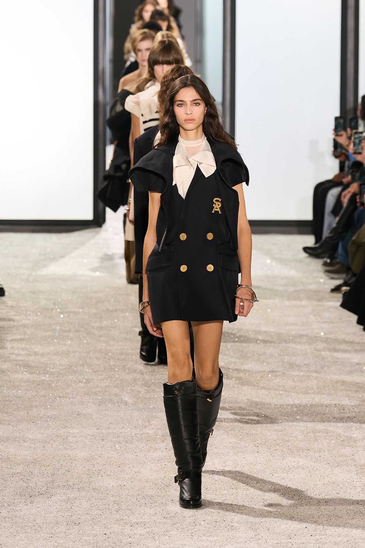Model walks the runway in a navy double-breasted coat-dress with gold buttons, beige blouse, and knee-high black boots, as other models line up behind her.