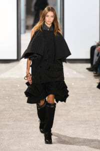 Model on a runway wearing a black pinstripe cape-style top with a tiered, ruffled skirt and tall black boots in a stark, minimalist setting.