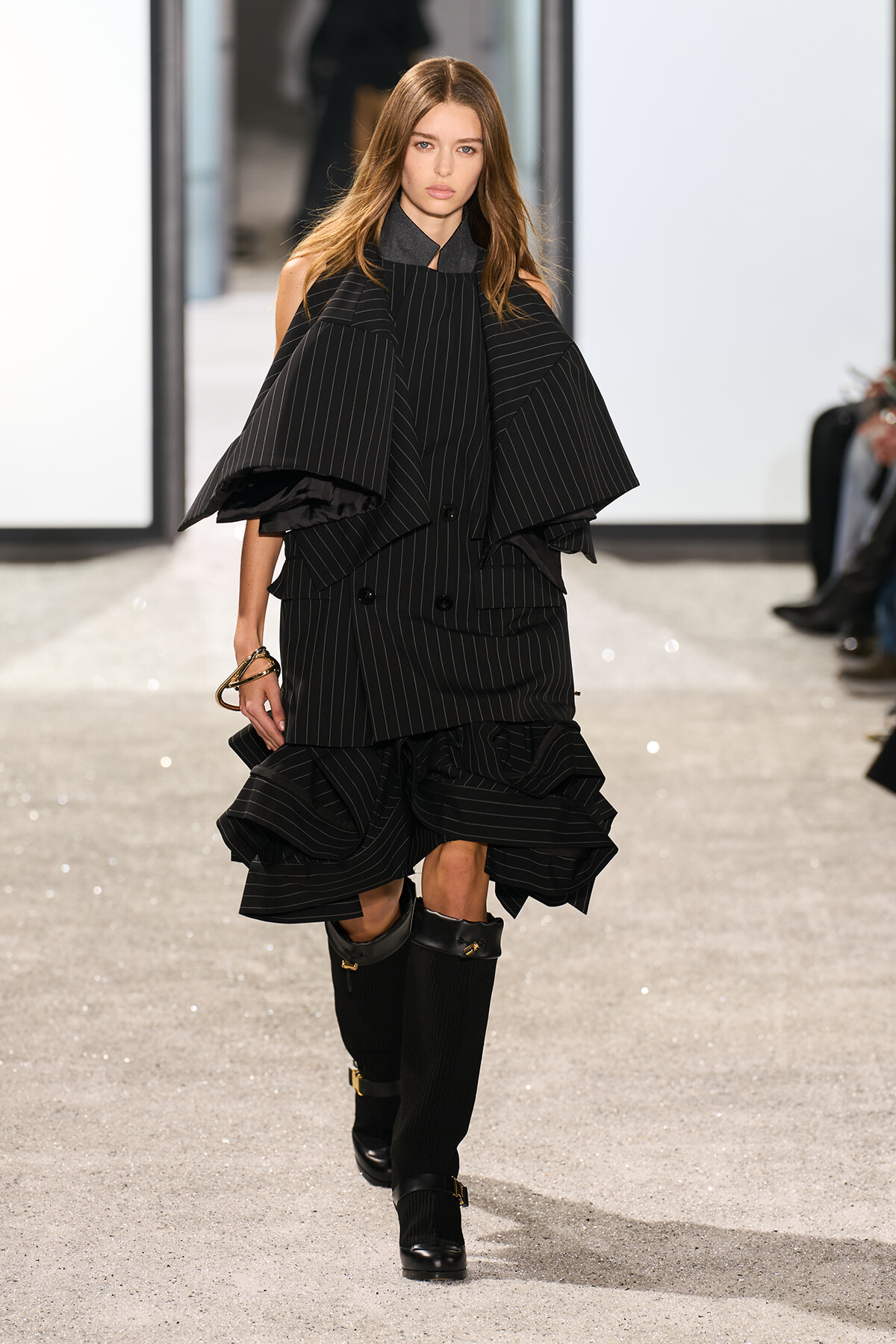 Model on a runway wearing a black pinstripe cape-style top with a tiered, ruffled skirt and tall black boots in a stark, minimalist setting.