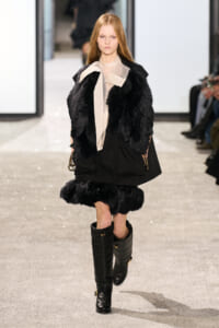 Model walking a fashion runway in a black fur vest over a beige blouse and black skirt, paired with tall black boots.