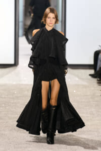 Model on a runway wearing a black pinstripe ensemble with voluminous layered cape sleeves and knee-high boots.